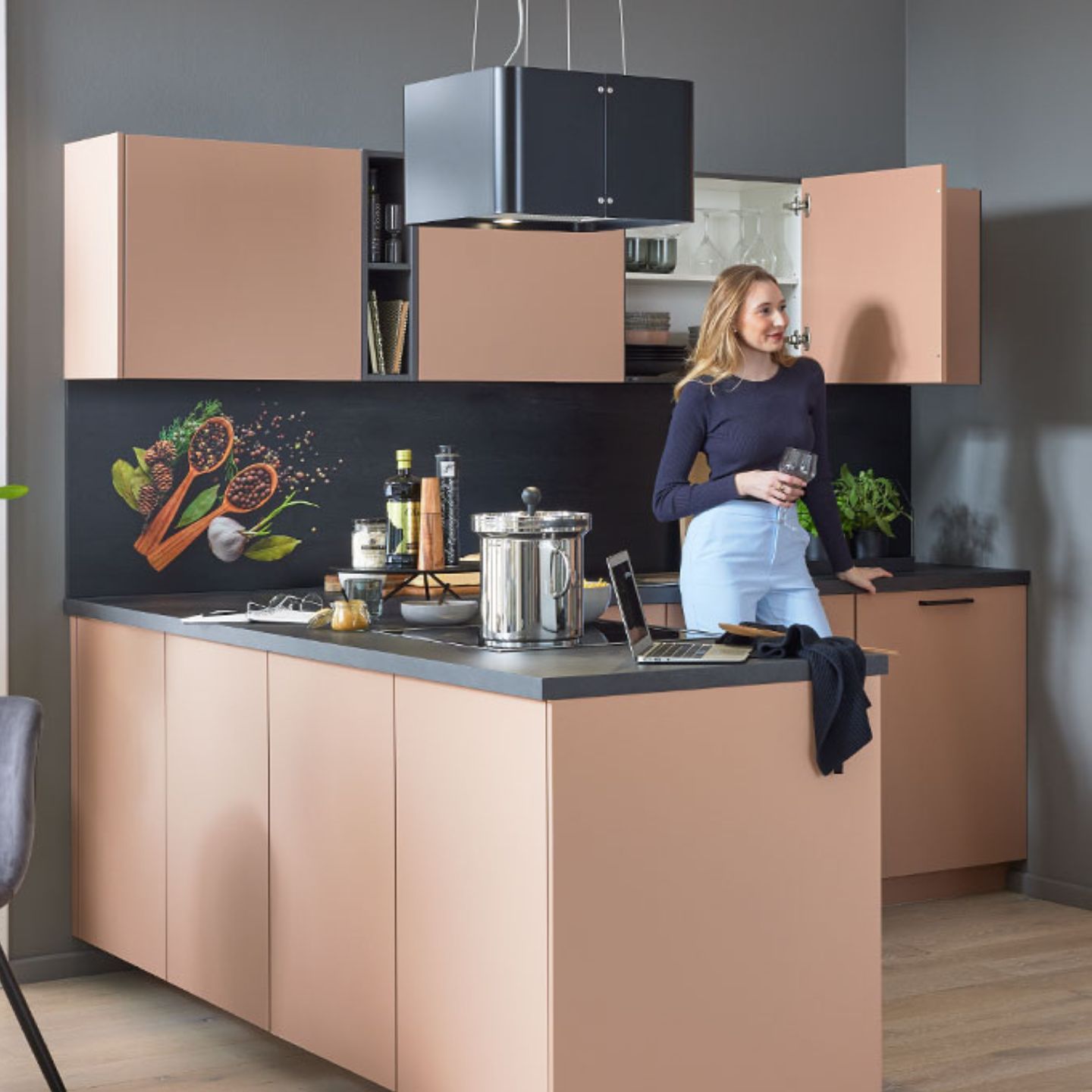 Eine Frau steht in einer modernen Küche mit pfirsichfarbenen Schränken und hält ein Glas Wein in der Hand. Auf dem Tresen stehen ein Laptop, Kochutensilien und ein Topf, auf der Arbeitsplatte sind Bilder von Gewürzen und Kräutern zu sehen. Eine Frau steht in einer modernen Küche mit pfirsichfarbenen Schränken und hält ein Glas Wein in der Hand. Auf dem Tresen stehen ein Laptop, Kochutensilien und ein Topf, auf der Arbeitsplatte sind Bilder von Gewürzen und Kräutern zu sehen.