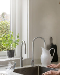 Eine Frau in einem gestreiften Kleid steht am Spülbecken und beobachtet, wie Dampf aus einem Quooker-Wasserhahn aufsteigt; im Bild sind ein Fenster, Pflanzen und Küchenutensilien zu sehen.