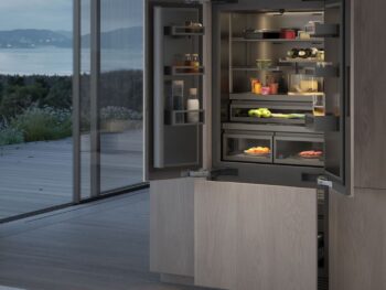 Gaggenau 400 Vario Serie: Ein moderner Einbaukühlschrank der Serie 400 von Gaggenau mit offenen Türen präsentiert ordentlich geordnete Lebensmittel und Getränke in einer Küche mit großen Fenstern und Blick auf einen malerischen Außenbereich.