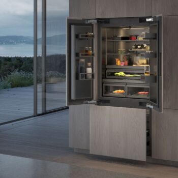 Gaggenau 400 Vario Serie: Ein moderner Einbaukühlschrank der Serie 400 von Gaggenau mit offenen Türen präsentiert ordentlich geordnete Lebensmittel und Getränke in einer Küche mit großen Fenstern und Blick auf einen malerischen Außenbereich.