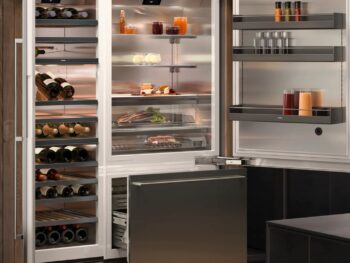 Gaggenau 400 Vario Serie: Offener moderner Kühlschrank von Gaggenau mit ordentlich angeordneten Weinflaschen, Getränken, Gewürzen und Lebensmitteln auf Ablagen, neben einer Gefrierschublade der Vario Serie 400, in einer modernen Küche.