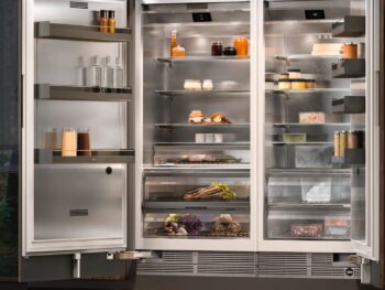Gaggenau 400 Vario Serie: Ein moderner Gaggenau 400 Doppeltür-Kühlschrank mit offenen Türen zeigt in einer Küche mit Holzvertäfelung auf Glasablagen ordentlich geordnete Lebensmittel, Getränke, Gewürze und Produkte.