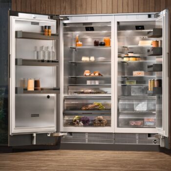 Gaggenau 400 Vario Serie: Ein moderner Gaggenau 400 Doppeltür-Kühlschrank mit offenen Türen zeigt in einer Küche mit Holzvertäfelung auf Glasablagen ordentlich geordnete Lebensmittel, Getränke, Gewürze und Produkte.