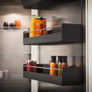 Gaggenau 400 Vario Serie: Zwei Ablagen in der Tür eines Gaggenau 400 Kühlschranks nehmen Flaschen und Gläser mit verschiedenen Flüssigkeiten und Konserven auf. Im Hintergrund sieht man das Innere des Kühlschranks, der zur 400 Vario Serie gehört, mit weiteren Gläsern.