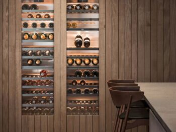 Gaggenau 400 Vario Serie: Eingebaute Weinkühler der Serie Gaggenau 400 Vario mit Glastüren stellen mehrere Flaschen auf Metallregalen aus, die in Holzvertäfelung neben einem Tresen mit Barhockern stehen.