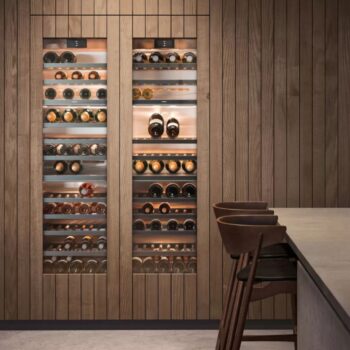 Gaggenau 400 Vario Serie: Eingebaute Weinkühler der Serie Gaggenau 400 Vario mit Glastüren stellen mehrere Flaschen auf Metallregalen aus, die in Holzvertäfelung neben einem Tresen mit Barhockern stehen.