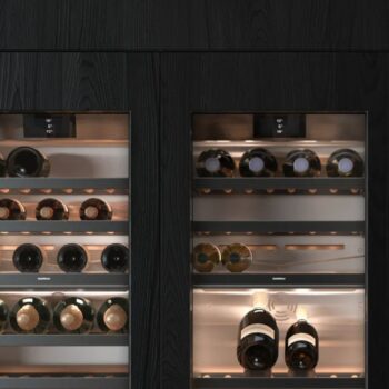 Gaggenau 400 Vario Serie: Zwei eingebaute Gaggenau 400 Vario Serie Weinkühlschränke mit Glastüren, die jeweils mehrere Weinflaschen enthalten, die horizontal auf Metallregalen gelagert sind, vor einem Hintergrund aus dunklem Holz.