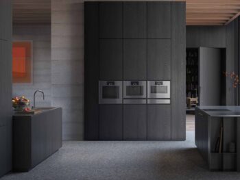 Gaggenau Expressive: Moderne Küche mit dunklen Schränken, Gaggenau-Edelstahl-Einbaubacköfen aus der Expressive Serie, einem kleinen Spülbereich und minimalistischem Dekor.