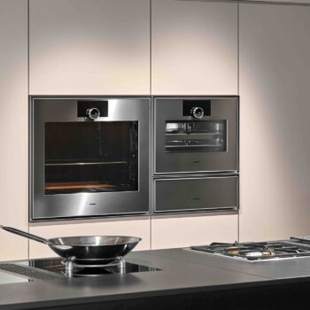 Gaggenau Expressive: Eine moderne Küche mit Gaggenau Edelstahl-Einbaubacköfen, einem Gas-Kochfeld und einer Bratpfanne auf der Theke im Vordergrund aus der Expressive Serie.