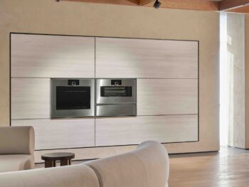 Gaggenau Expressive: Modernes Wohnzimmer mit eingebauten Gaggenau Edelstahlöfen der Expressive Serie in einer hellen holzgetäfelten Wand, beigem Sofa im Vordergrund und Holzfußboden.