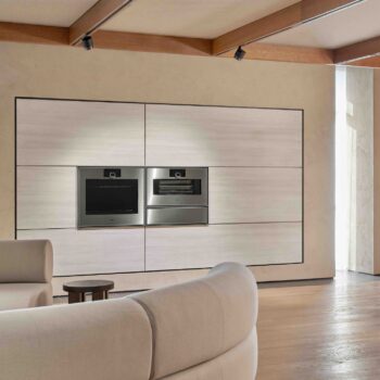 Gaggenau Expressive: Modernes Wohnzimmer mit eingebauten Gaggenau Edelstahlöfen der Expressive Serie in einer hellen holzgetäfelten Wand, beigem Sofa im Vordergrund und Holzfußboden.