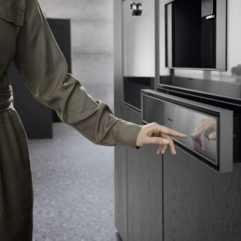 Gaggenau Expressive: Eine Person in grünem Outfit berührt die Vorderseite eines modernen Kücheneinbaugeräts der Gaggenau Expressive Serie mit einer reflektierenden Oberfläche, möglicherweise ein Backofen oder eine Mikrowelle.