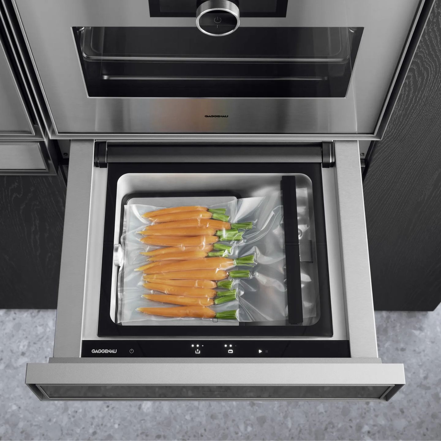 Ein Vakuumiergerät der Gaggenau Expressive Serie mit vakuumversiegelten frischen Karotten in einem Plastikbeutel unter einem Einbauofen in einer modernen Küche.