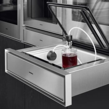 Gaggenau Expressive: Eine Gaggenau Edelstahl-Vakuumierschublade mit offenem Deckel aus der Expressive Serie vakuumiert mit einem angeschlossenen Schlauch eine Glasflasche mit einer dunklen Flüssigkeit.