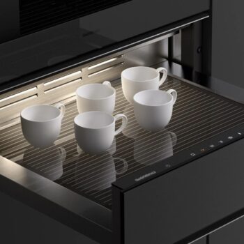 Gaggenau Expressive: Fünf weiße Keramiktassen aus der Expressive Serie stehen auf einer gestreiften Gaggenau Wärmeschublade unter einer Einbauleuchte in einer modernen Küche.