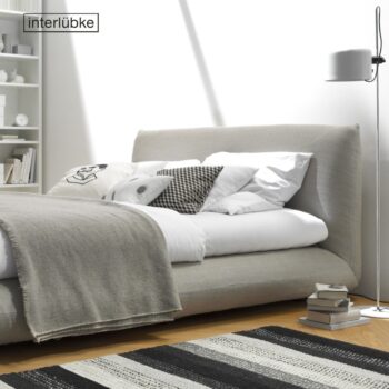 Ein modernes Schlafzimmer mit einem niedrigen, gepolsterten Bett Jalis von Interlübke, neutraler Bettwäsche, einer Stehlampe, gestapelten Büchern und einem gestreiften Teppich auf einem Holzboden. Im Hintergrund sind Regale mit Büchern und Dekoration zu sehen.