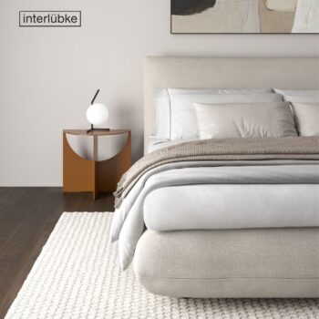 Minimalistisches Schlafzimmer mit einem beigen Interlübke-Bett, geschichtetem Bettzeug, einem braunen Beistelltisch mit einer runden Lampe, einem weißen Teppich und abstrakter Wandkunst. Das von Jalis inspirierte Interlübke-Logo ist an der Wand zu sehen.