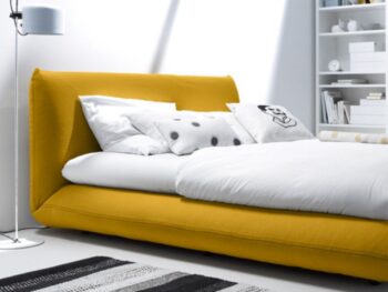 Ein modernes Schlafzimmer mit einem niedrigen gelben Bett Jalis, weißen und gemusterten Kissen, einem monochromen Teppich, einer Stehlampe und eingebauten Regalen. Das Wort interlübke erscheint oben links.