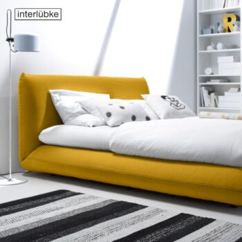 Ein modernes Schlafzimmer mit einem niedrigen gelben Bett Jalis, weißen und gemusterten Kissen, einem monochromen Teppich, einer Stehlampe und eingebauten Regalen. Das Wort interlübke erscheint oben links.