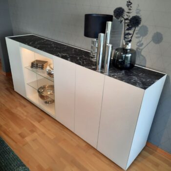 Modernes weißes Interlübke-Sideboard mit schwarzer Marmorplatte, Glasböden mit Dekorationsartikeln, dekorativen Vasen und einer Lampe auf der Oberfläche, an einer gemusterten Wand platziert.