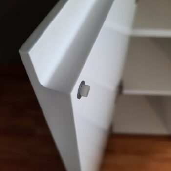 Nahaufnahme eines Interlübke cube light Sideboards, weißes Regalpaneel mit seitlich eingefügten Regalstützen aus Metall und Kunststoff; im Hintergrund sind Regale und Holzboden zu sehen.