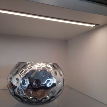 Eine spiegelnde silberne Schale mit kreisrunden Vertiefungen wird auf einer Glasablage unter einem LED-Lichtband präsentiert und ergänzt elegant das Interlübke cube light Sideboard.