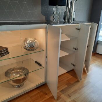 Ein weißes Sideboard in Würfelform mit zwei offenen Türen gibt den Blick auf leere Fächer frei; in der angrenzenden Glasvitrine befinden sich zwei dekorative Metallschalen und eine Fernbedienung.