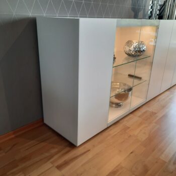 Ein moderner weißer Interlübke-Schrank mit Glastüren zeigt silberne Dekoschalen auf Glasböden, die auf einem Holzboden vor einer gemusterten grauen Wand stehen - ein elegantes, leichtes Würfel-Sideboard für moderne Räume.