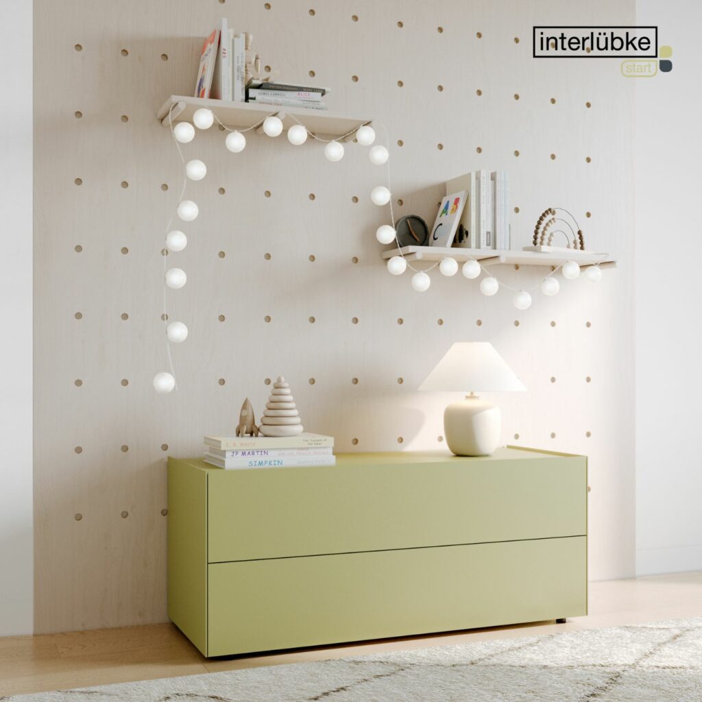 Eine hellgrüne Interlübke-Kommode mit Lampe und Dekoration steht an einer Wand mit Lochplatten; zwei weiße Regale über der Anrichte beherbergen Bücher, Ornamente und Lichterketten.
