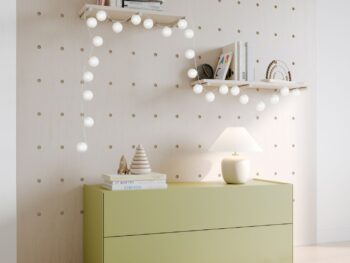 Eine hellgrüne Interlübke-Kommode mit Lampe und Dekoration steht an einer Wand mit Lochplatten; zwei weiße Regale über der Anrichte beherbergen Bücher, Ornamente und Lichterketten.