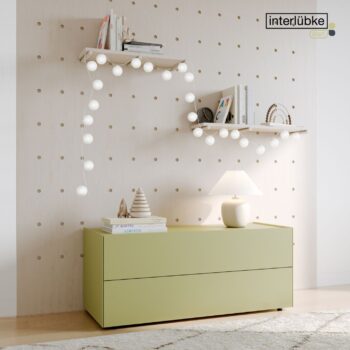 Eine hellgrüne Interlübke-Kommode mit Lampe und Dekoration steht an einer Wand mit Lochplatten; zwei weiße Regale über der Anrichte beherbergen Bücher, Ornamente und Lichterketten.