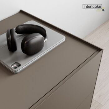 Ein Paar Over-Ear-Kopfhörer und ein Smartphone ruhen auf einem geschlossenen Laptop, der auf einem mattbraunen Sideboard steht. Das Interlübke-Logo erscheint in der oberen rechten Ecke und sorgt für einen perfekten Start in den Tag.