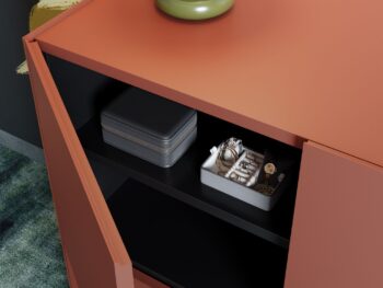 Ein orangefarbenes Interlübke-Sideboard mit offenen Türen gibt den Blick frei auf ein graues Kästchen und ein Tablett mit Schmuck und einer Uhr in einem schwarzen Regal.