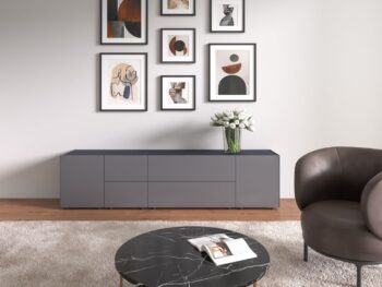 Modernes Wohnzimmer mit einem Interlübke-Sideboard, einer Wand mit abstrakter Kunst, einem runden Couchtisch aus schwarzem Marmor, einem Sessel und einer Vase mit weißen Blumen, die von einer eleganten Würfelleuchte beleuchtet wird.