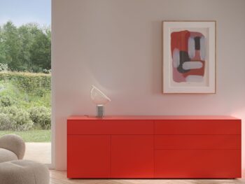 Ein modernes Zimmer mit einem Interlübke Sideboard in Rot, einem beigen strukturierten Sessel, abstrakten gerahmten Kunstwerken an der Wand und einer Glastür, die auf einen grünen Garten blickt, der von sanftem Cube-Licht beleuchtet wird.