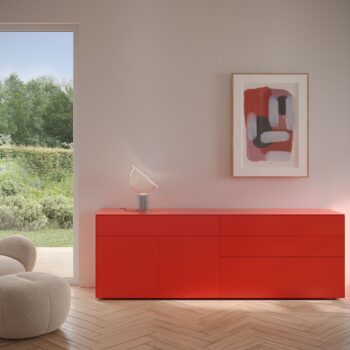 Ein modernes Zimmer mit einem Interlübke Sideboard in Rot, einem beigen strukturierten Sessel, abstrakten gerahmten Kunstwerken an der Wand und einer Glastür, die auf einen grünen Garten blickt, der von sanftem Cube-Licht beleuchtet wird.
