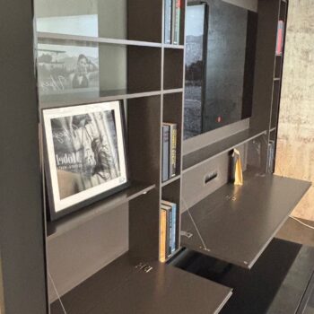 Ein modernes dunkles Interlübke-Regal mit Büchern, gerahmten Schwarz-Weiß-Fotografien und zwei ausklappbaren Schreibtischflächen vor einer Betonwand - perfekt als stilvolles TV-Wandregal oder für einen bücherlosen, minimalistischen Look.