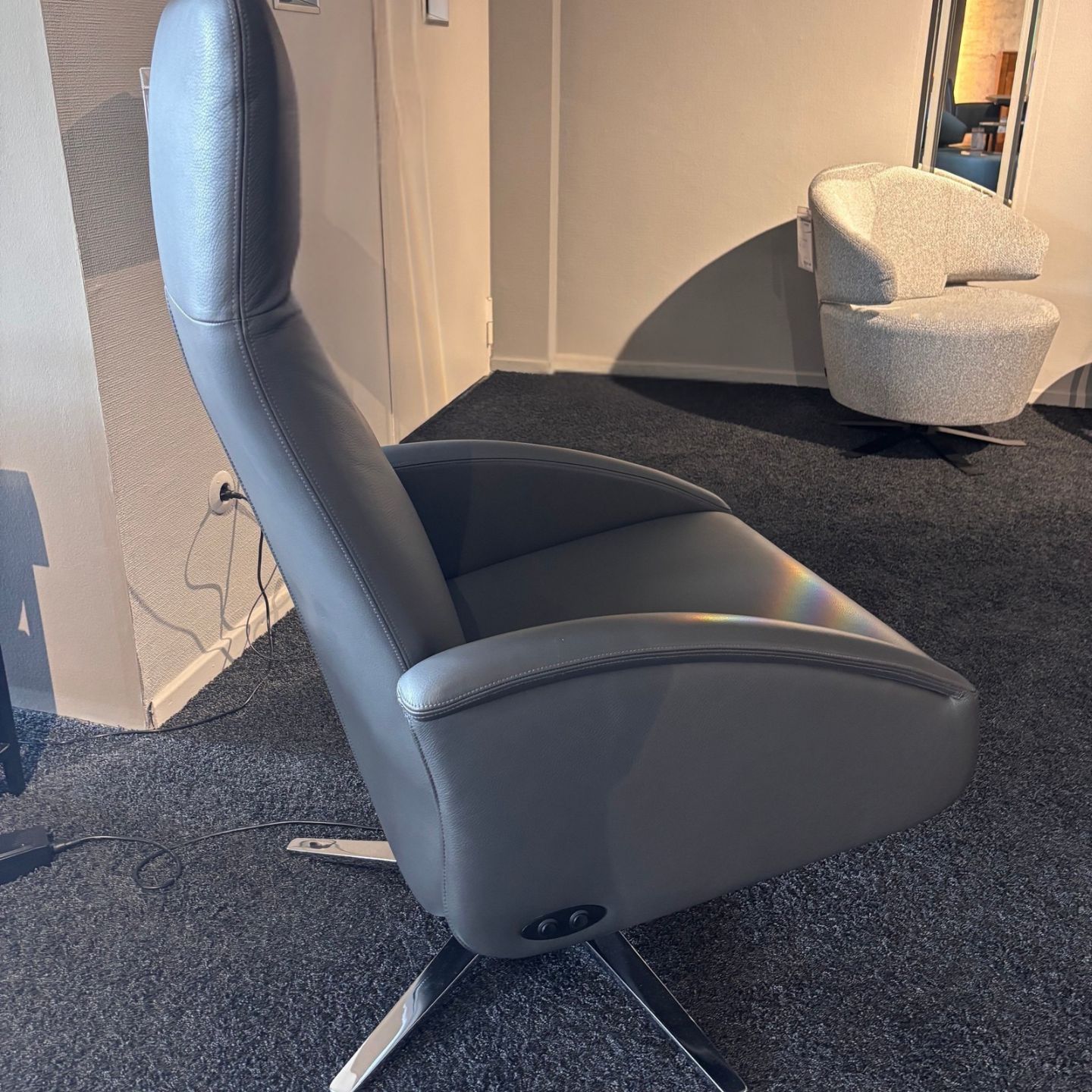 Im Cliff-Showroom steht ein moderner grauer Funktionssessel mit Motor, hoher Rückenlehne und Metallbeinen auf einem dunklen Teppich. Im Hintergrund ist ein zweiter, hellerer Sessel zu sehen. Im Cliff-Showroom steht ein moderner grauer Funktionssessel mit Motor, hoher Rückenlehne und Metallbeinen auf einem dunklen Teppich. Im Hintergrund ist ein zweiter, hellerer Sessel zu sehen.