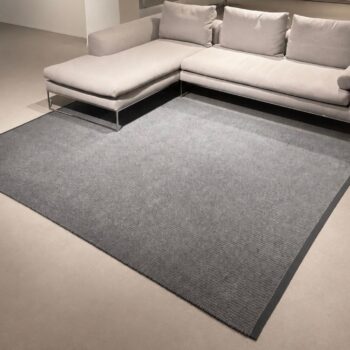 Ein modernes, hellgraues Ecksofa steht auf einem großen, rechteckigen Kasthall-Designteppich in einem minimalistisch gestalteten Raum mit beigem Teppichboden und neutralen Wänden.