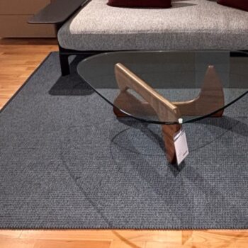 Ein Couchtisch aus Glas mit Holzbeinen steht auf einem grauen Teppich von Kasthall Designteppich vor einem modernen grauen Sofa mit zwei weinroten Kissen auf einem hellen Holzboden.