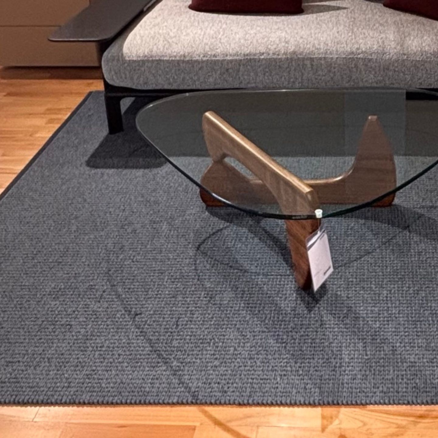 Ein Couchtisch aus Glas mit Holzbeinen steht auf einem grauen Teppich von Kasthall Designteppich vor einem modernen grauen Sofa mit zwei weinroten Kissen auf einem hellen Holzboden. Ein Couchtisch aus Glas mit Holzbeinen steht auf einem grauen Teppich von Kasthall Designteppich vor einem modernen grauen Sofa mit zwei weinroten Kissen auf einem hellen Holzboden.