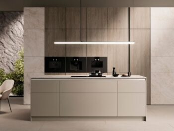 Moderne, minimalistische Küche mit zentraler Insel, Einbaugeräten an einer holzgetäfelten Wand und einer linearen Pendelleuchte; im Hintergrund ist die Grünanlage zu sehen.