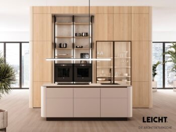 Moderne Küche mit hellen Holzschränken, einer zentralen Insel, eingebauten Öfen, offenen Regalen mit Küchenutensilien und Glasschränken. Große Fenster sorgen für reichlich natürliches Licht.