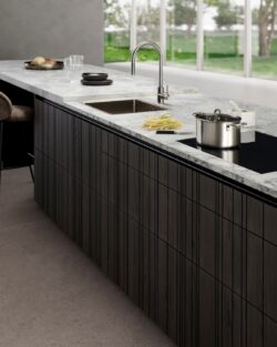 Eine moderne Bahia LEICHT-Küche mit Marmorarbeitsplatte, Einbauspüle, Induktionskochfeld, Topf, geschnittenen Kartoffeln und einem Stuhl, mit großen Fenstern im Hintergrund.