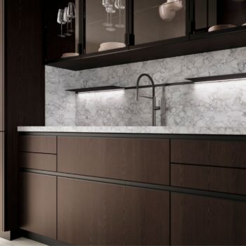 Moderne Küche der Bahia Serie mit Schränken aus dunklem Holz, Aufkantung und Arbeitsplatte aus Marmor, Unterbaubeleuchtung sowie eingebautem Wasserhahn und Spülbecken. Obere Glasschränke von LEICHT bieten Platz für Geschirr und Gläser und verleihen der Küche eine von Bahia inspirierte Eleganz.
