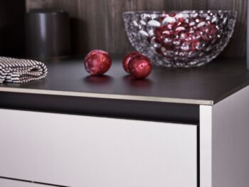 Leicht Küchen Contino E12 - Eine Arbeitsplatte von Leicht Küchen zeigt eine mit roten Ornamenten gefüllte, strukturierte Glasschale, daneben zwei rote Kugeln, ein gefaltetes, gestreiftes Tuch und im Hintergrund einen dunklen, zylindrischen Behälter - eine perfekte Ergänzung für Contino E12-Küchen.