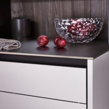 Leicht Küchen Contino E12 - Eine Arbeitsplatte von Leicht Küchen zeigt eine mit roten Ornamenten gefüllte, strukturierte Glasschale, daneben zwei rote Kugeln, ein gefaltetes, gestreiftes Tuch und im Hintergrund einen dunklen, zylindrischen Behälter - eine perfekte Ergänzung für Contino E12-Küchen.