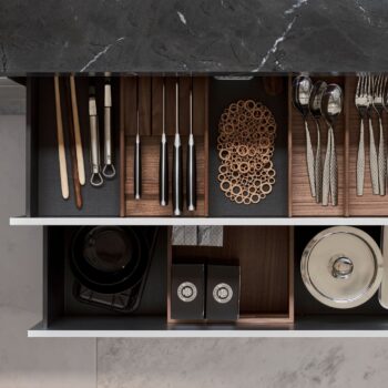 Leicht Küchen Contino E12 - Offene Küchenschublade mit Utensilien, Essstäbchen, Messern, Metalluntersetzern, Schüsseln, Tellern und schwarzen Behältern mit Deckeln unter einer Arbeitsplatte aus Marmor, die das elegante Design von Leicht Küchen unterstreicht.