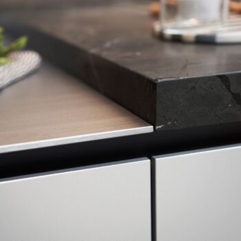 Leicht Küchen Contino E12 - Die Nahaufnahme einer Küchenarbeitsplatte zeigt den Kontrast zwischen einer dunklen Steinoberfläche und einem Leicht Küchen Contino E12 Schrank aus gebürstetem Metall darunter.