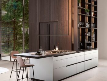 Leicht Küchen Contino E12 - Moderne Küche mit großer Insel, eingebautem Herd, Spüle, zwei Barhockern, offenen Regalen und hohen Holzschränken, mit großen Fenstern, die den Blick ins Grüne freigeben.